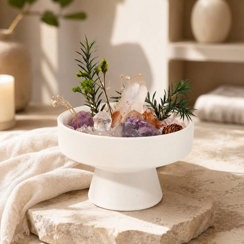 White bowl on pedestal with greenery, pinecones, and pink, purple, and clear crystals