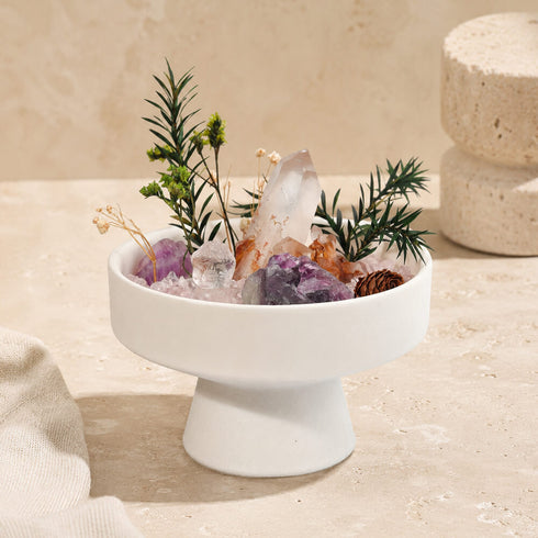 White crystal bowl centerpiece with green sprigs, pinecones, and pink and purple crystal pieces